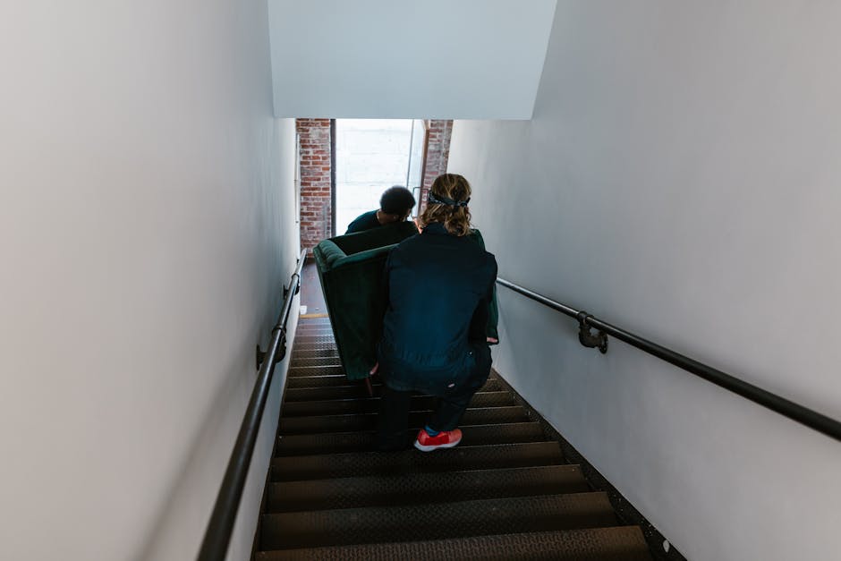 Two men carefully move a couch down a staircase in an indoor setting.